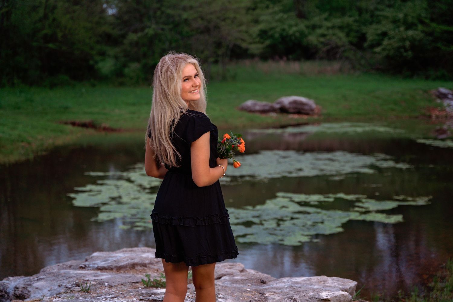 Person in a black dress stands near a pond with lily pads at dusk holding flowers in a tranquil outdoor setting.