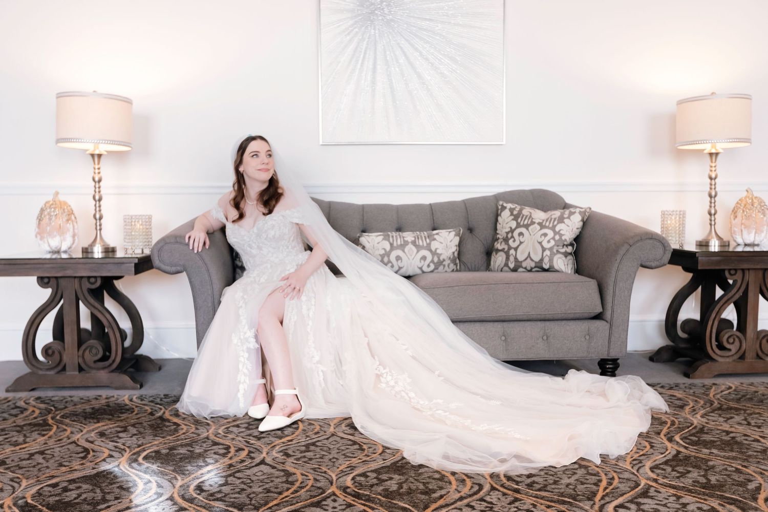 Elegant bride in lace off-shoulder wedding gown sitting gracefully on a gray sofa in a luxury venue.