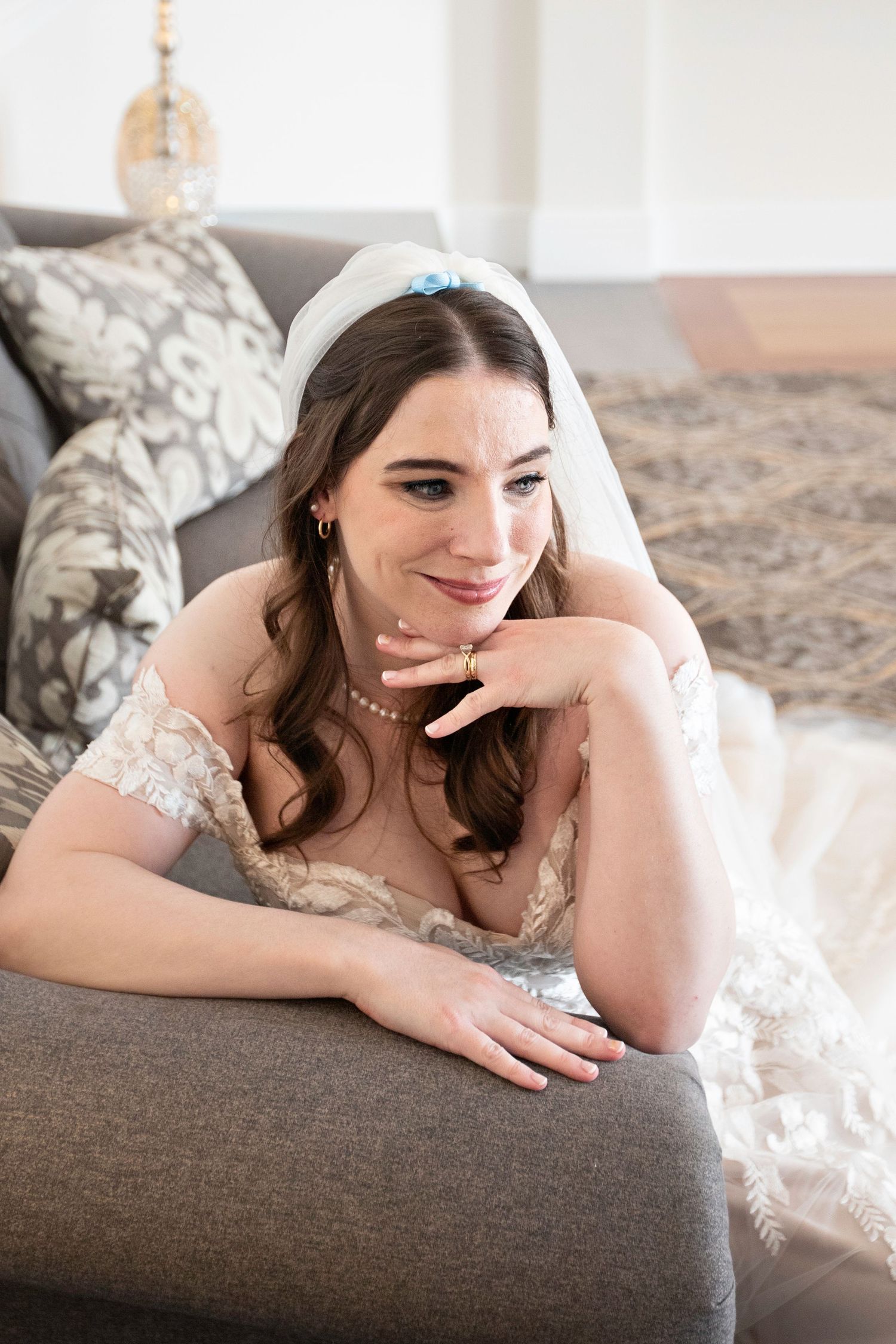 Beautiful bride in lace wedding dress and veil resting on a gray sofa with a soft smile.