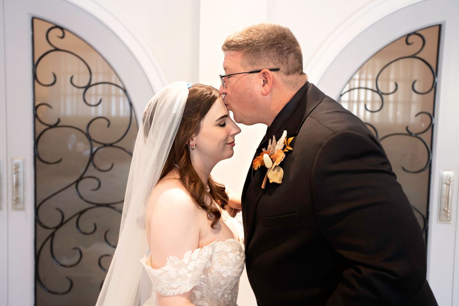 Groom tenderly kissing bride near ornate arched double doors during wedding ceremony.