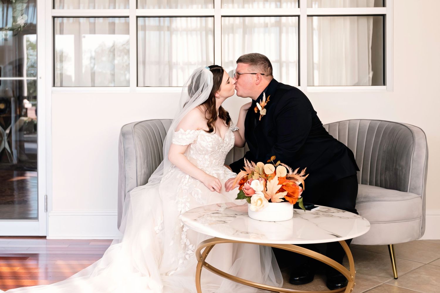 Bride and groom sharing a romantic kiss while seated together with a floral bouquet on a marble table.