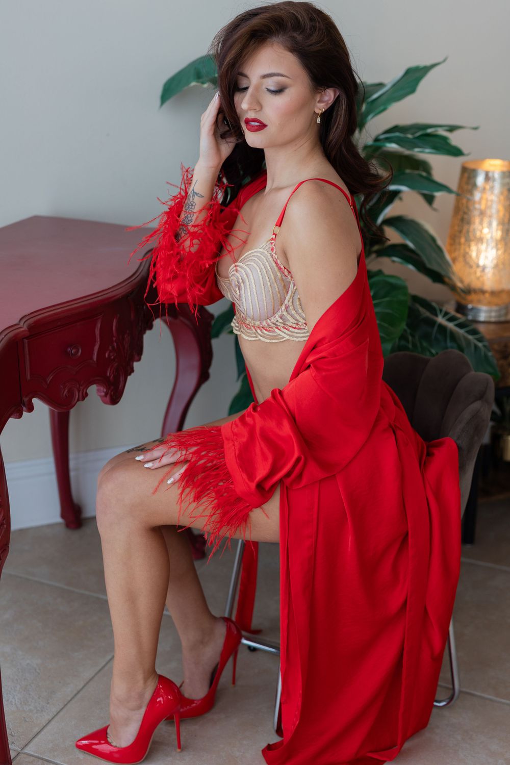 Glamorous boudoir photo featuring beige lingerie, red feather robe, and matching red high heels in an elegant setting.