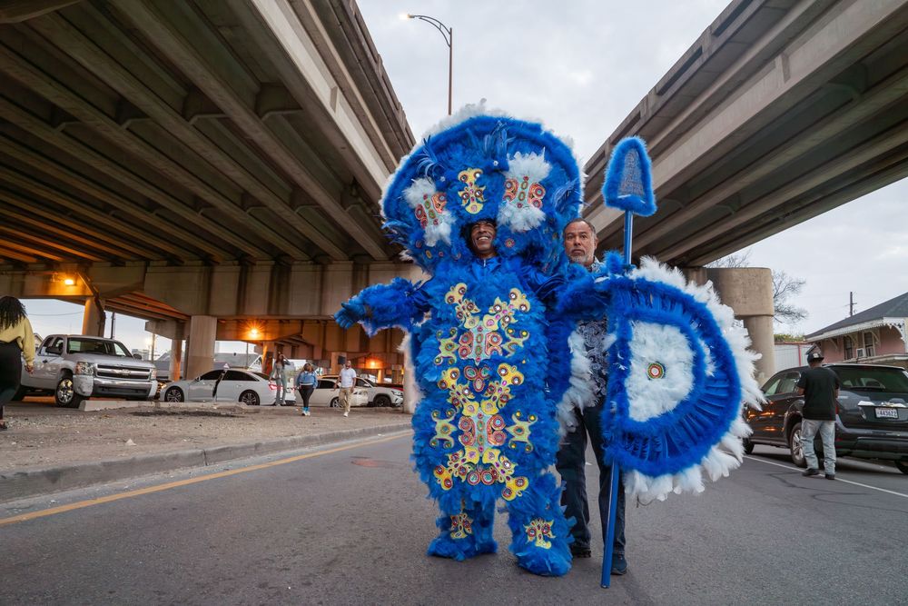 A performer in a bright blue feathered carnival costume dances beneath a highway overpass during a street performance.