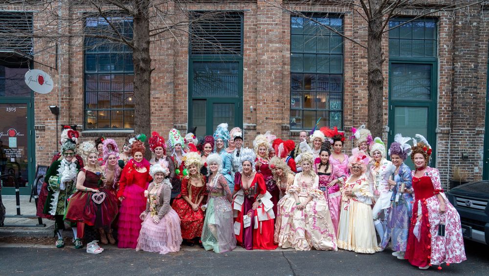 A group of people in elaborate period costumes and dresses pose together outside a brick building.