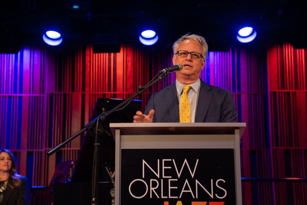 Speakers stand at podiums with colorful stage lighting and 'New Orleans' text visible in background during event presentation.