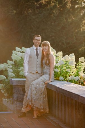 Couple poses on stone balcony with blooming white hydrangeas in golden sunset light.