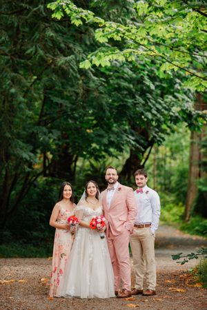 Wedding party of four posing on a forest path, bride in white gown with red bouquet, groom in pink suit.