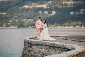 Bride and groom posing on stone waterfront pier with mountain lake backdrop, groom in pink suit, bride in white gown.