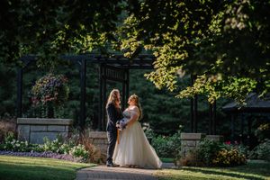 Two people celebrate their wedding in a beautiful garden setting with a floral gazebo and late afternoon sunlight.