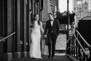 An elegant couple walks hand in hand along an urban sidewalk in formal wedding attire.
