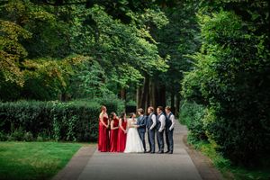A wedding party in red and gray attire walks along a tree-lined path in a lush garden setting.