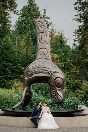 Wedding couple poses in front of large curved First Nations art sculpture in Pacific Northwest garden setting.