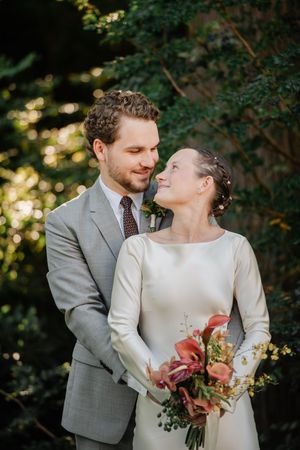 A couple shares a tender moment outdoors in wedding attire with a grey suit and simple white dress holding a pink bouquet.