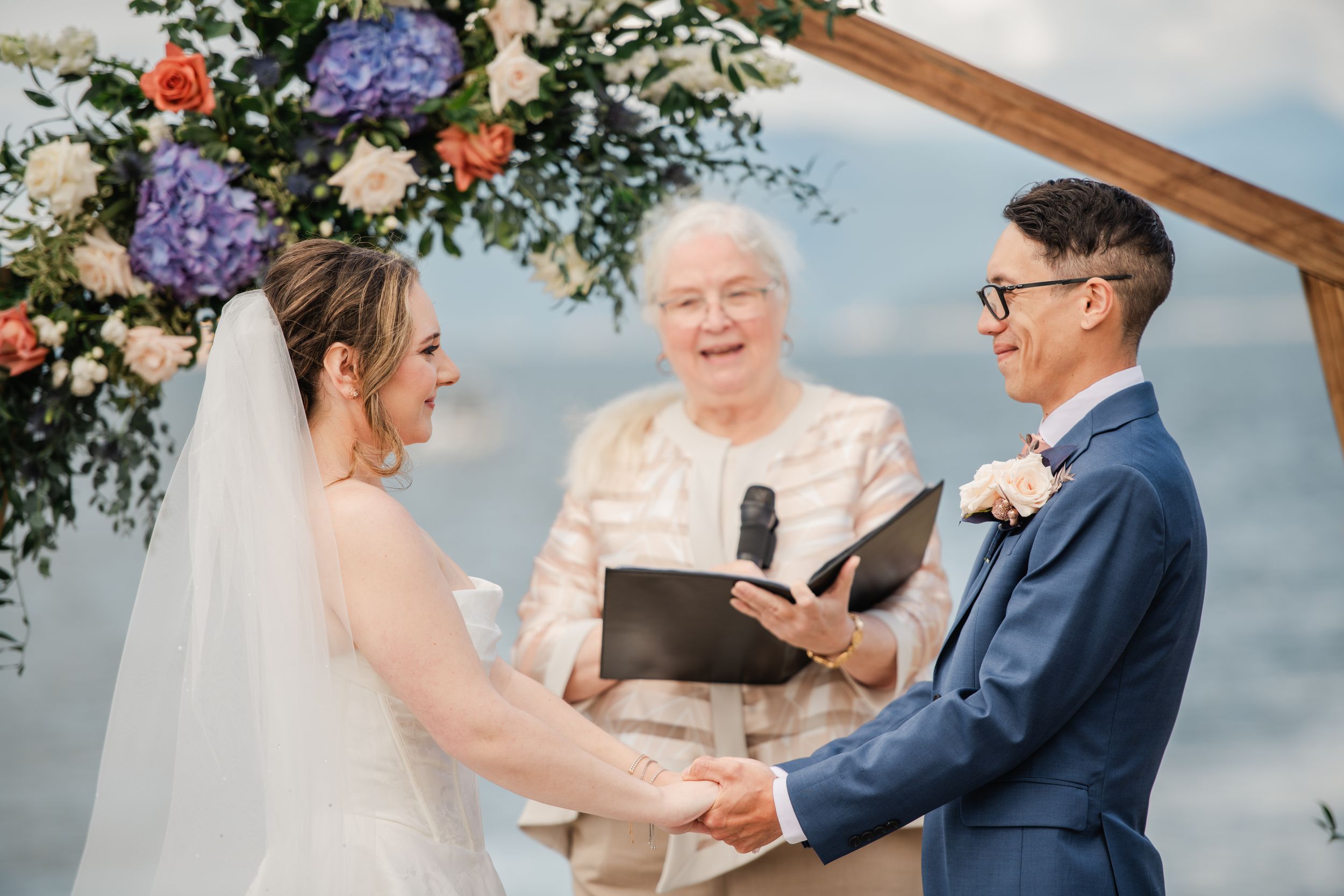 Bride and groom exchange vows outdoors under floral arch with officiant, waterfront wedding ceremony.