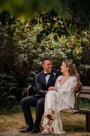 A couple shares a joyful moment while sitting on a garden bench in wedding attire.