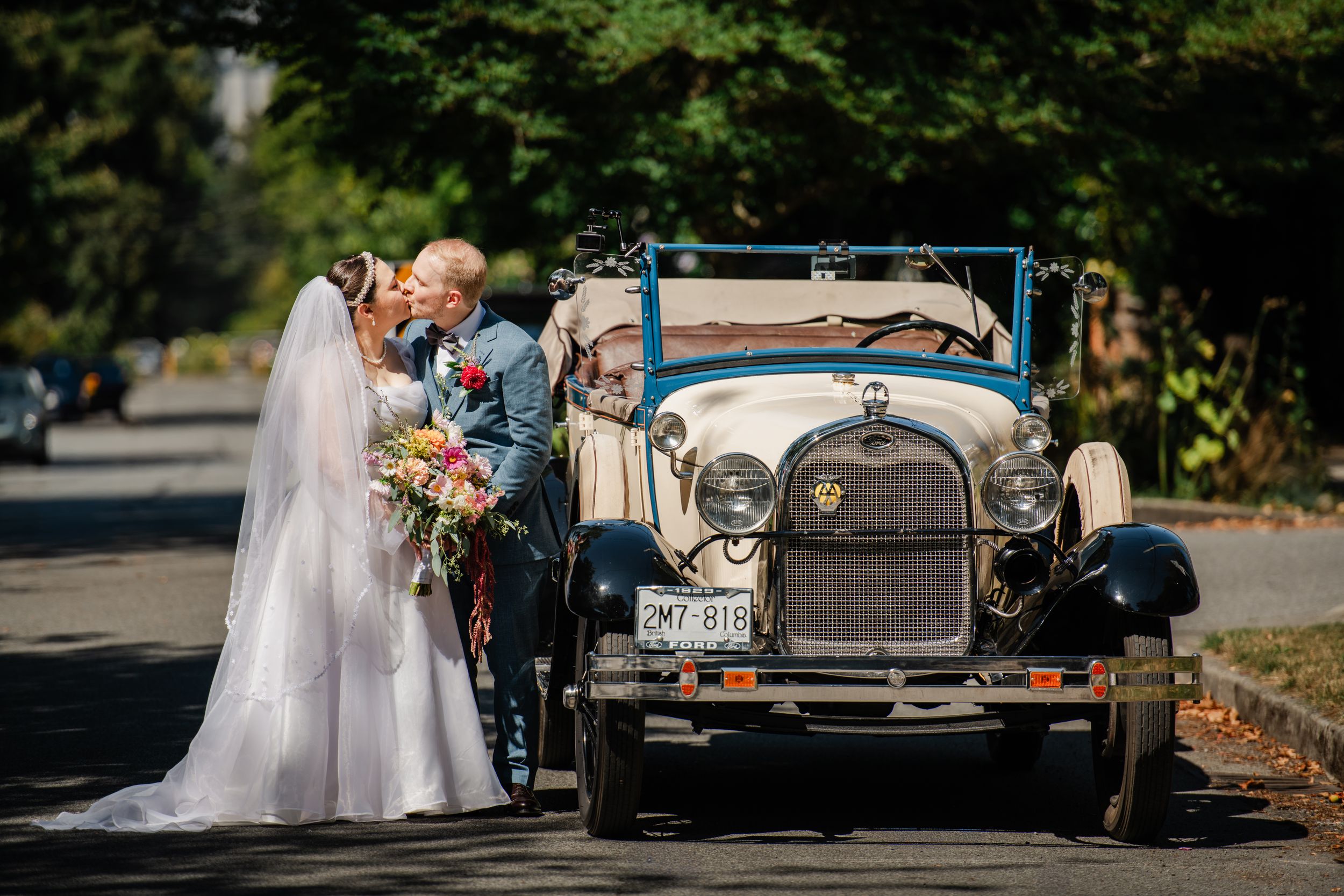Bride and groom kissing beside a vintage classic car on their wedding day, surrounded by lush green trees.