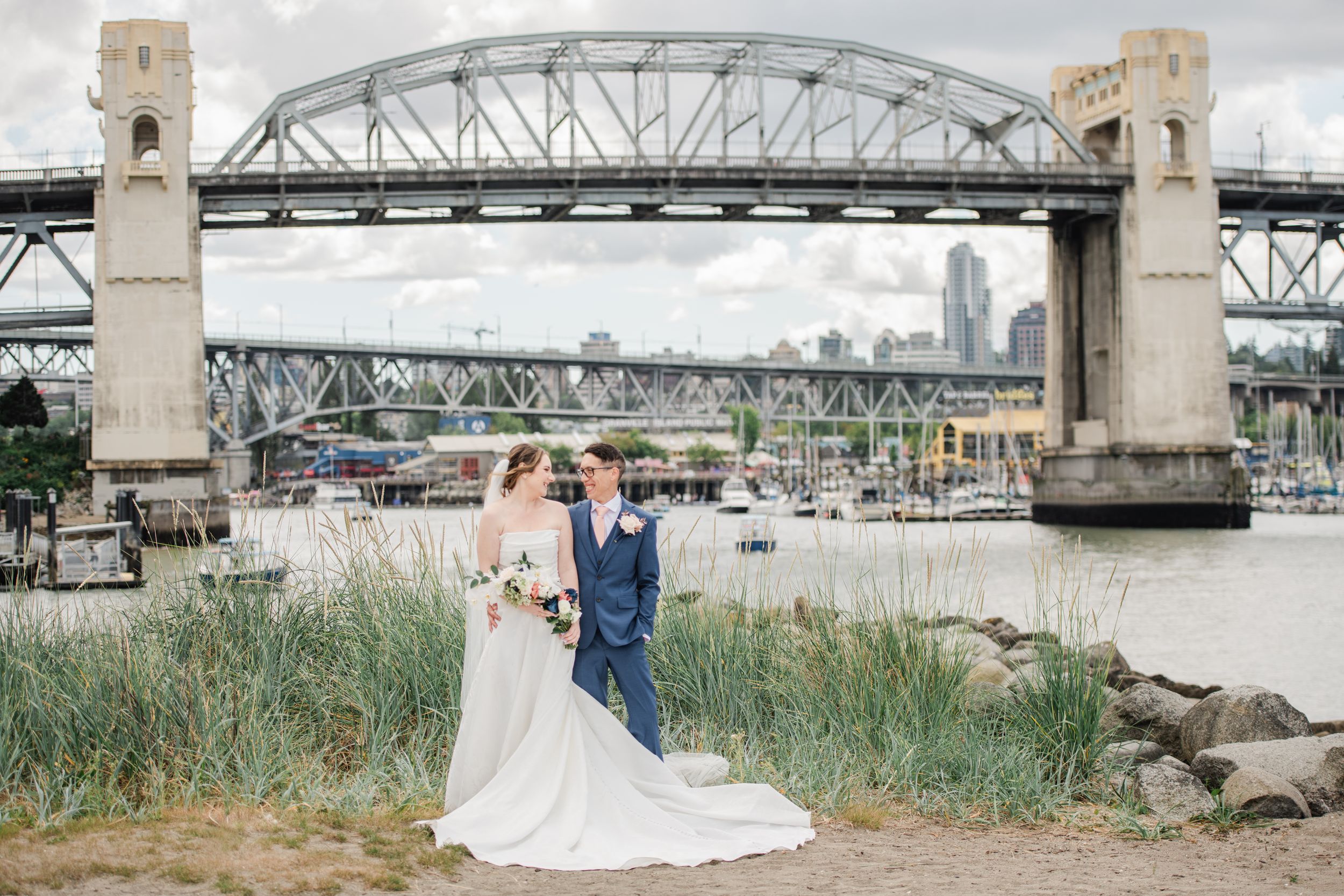Bride and groom pose near Burrard Bridge in Vancouver with marina and city skyline in the background.