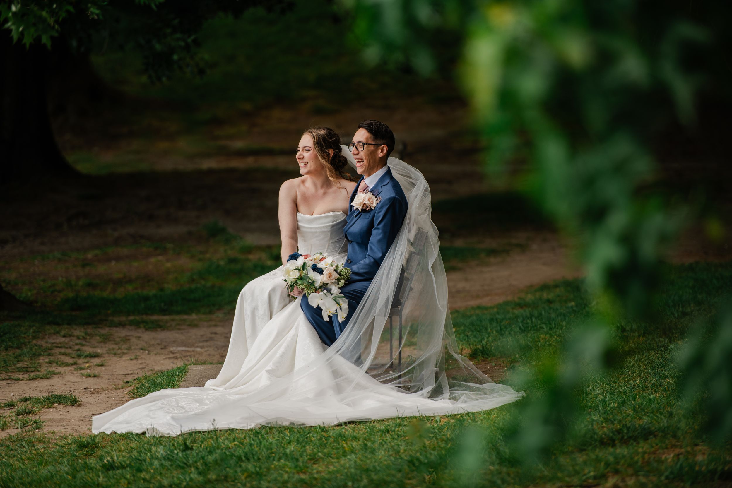 Bride and groom sit together on grass surrounded by lush green trees in a romantic outdoor wedding portrait.