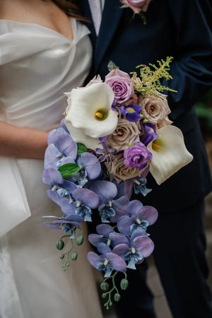 Close-up view of elegant wedding bouquet with white calla lilies, purple hydrangeas, and mauve roses against formal attire.