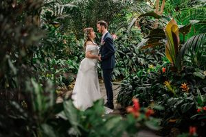 Two newlyweds share an intimate moment surrounded by lush tropical plants and flowers in a greenhouse setting.