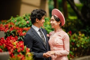 Couple in traditional Vietnamese ao dai and suit smiling at each other outdoors with red flowers.