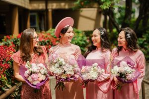 Four women in pink ao dai dresses holding bouquets of flowers, smiling outdoors at a Vietnamese wedding.