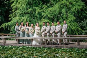 Wedding party wearing gray and white attire poses together on a rustic bridge surrounded by lush green foliage.