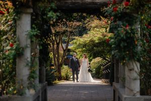 A couple walks through a garden archway adorned with climbing vines and red flowers.