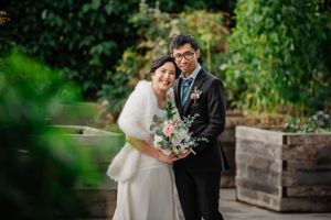 A couple in wedding attire embraces outdoors while holding a bouquet in a garden setting with stone walls.