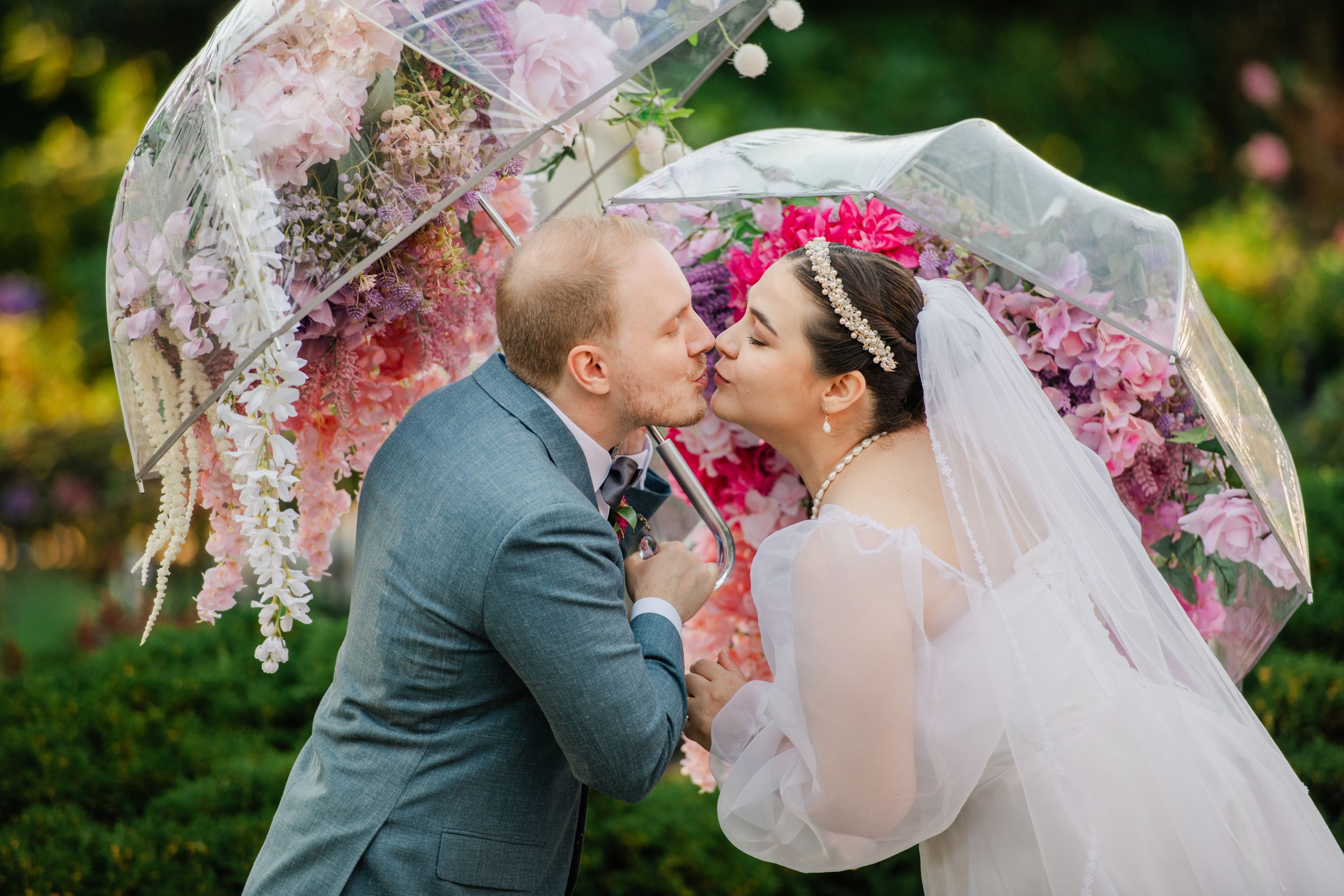 Bride and groom kissing under a floral decorated clear umbrella in a lush garden on their wedding day.