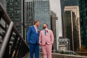 A couple in coordinating pink and blue suits poses on a rooftop with city buildings in background.