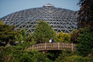 A couple poses on a wooden bridge with a large geodesic dome structure visible in the background.
