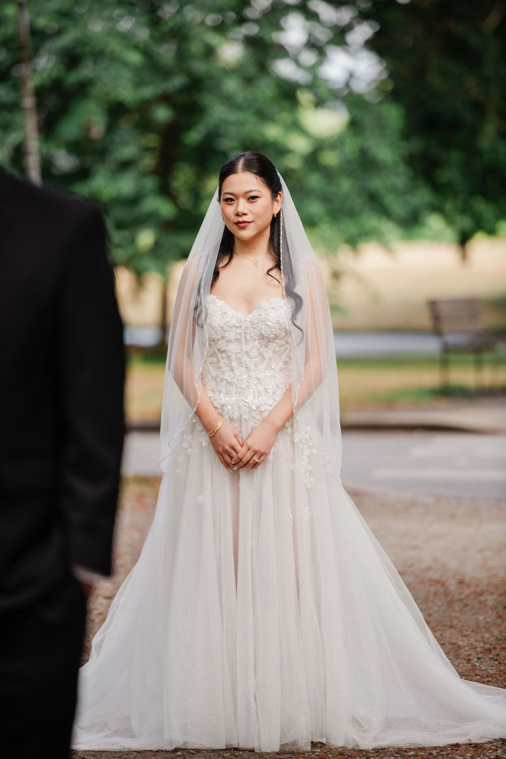 Bride in a lace strapless wedding gown with veil stands in a park, facing groom in black suit.