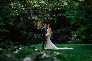 Newlyweds share intimate moment in sunlit garden clearing surrounded by lush green foliage.
