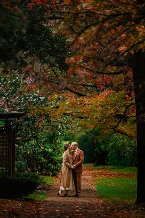A couple walking together on a leaf-covered path beneath vibrant autumn trees in a park setting.