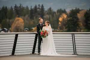 A couple in wedding attire stands together on a deck overlooking evergreen trees and autumn foliage.