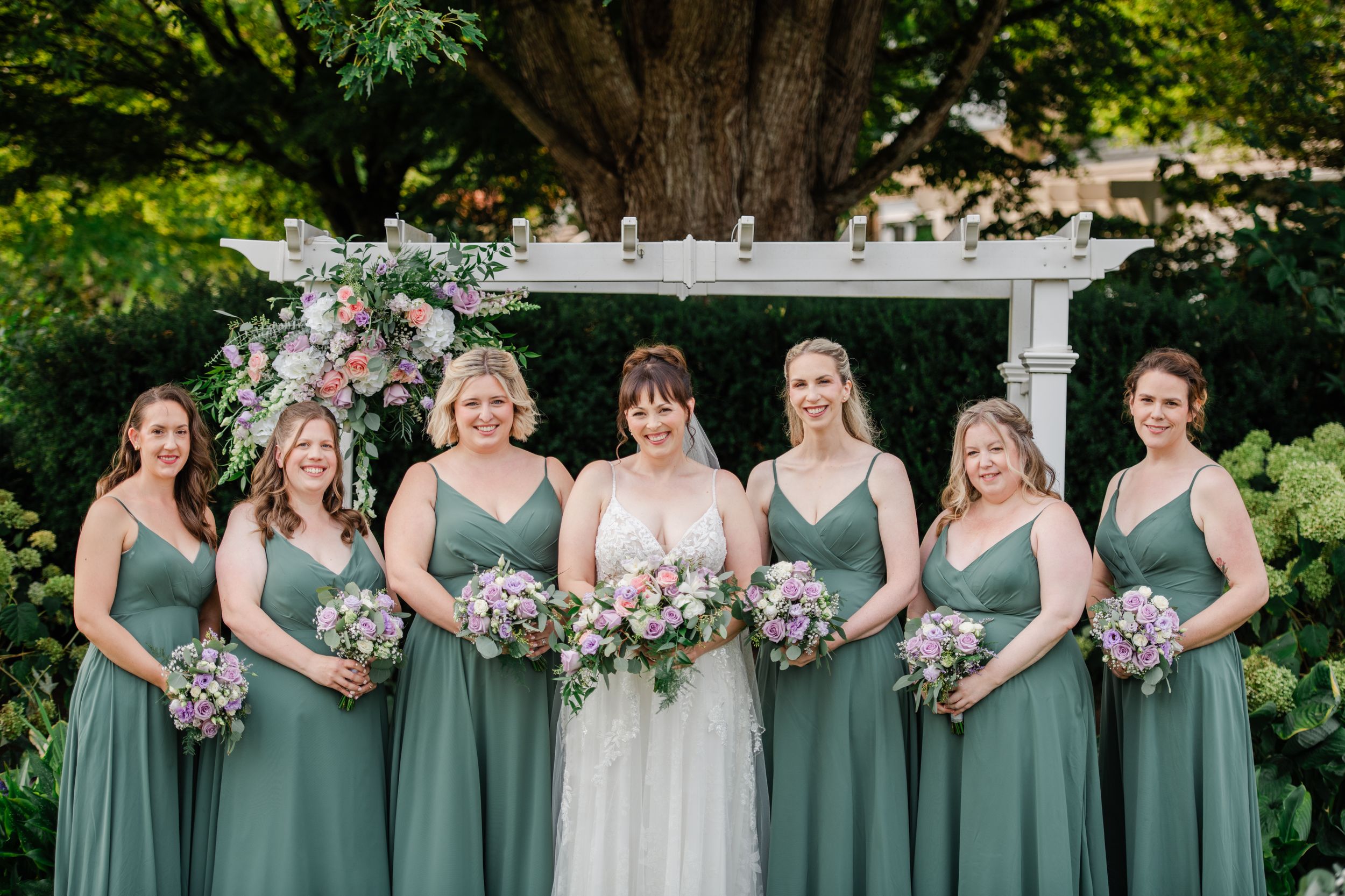 Bride in white gown with six bridesmaids in sage green dresses holding pink bouquets outdoors at a wedding ceremony.