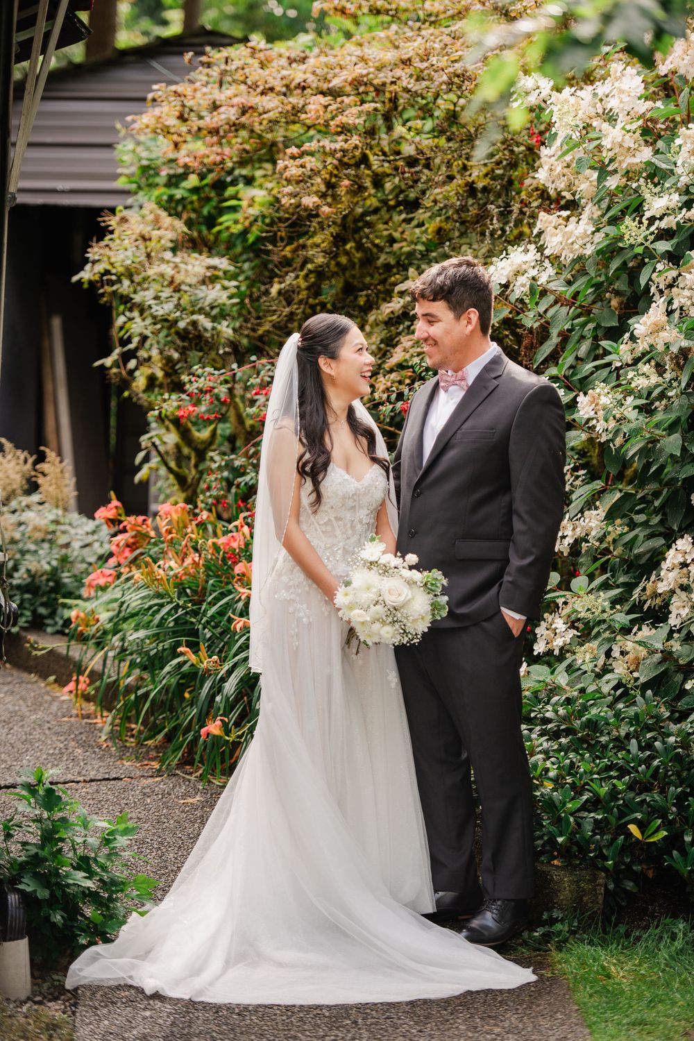 Bride and groom stand together in a lush garden, she holds white bouquet, wearing gown with veil.