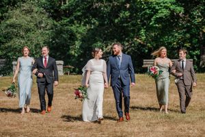 Wedding party in formal attire walks together across a grassy outdoor field during summer festivities.