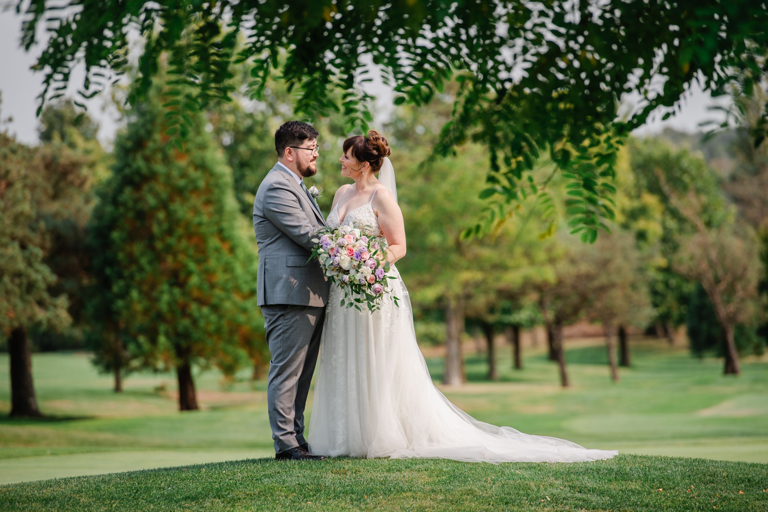 Bride and groom share a romantic moment on lush green golf course grounds, surrounded by trees, bride holding floral bouquet.