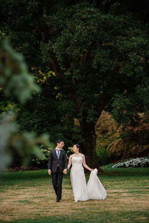 A couple takes a romantic stroll on a lush green lawn surrounded by trees at an elegant outdoor garden venue.