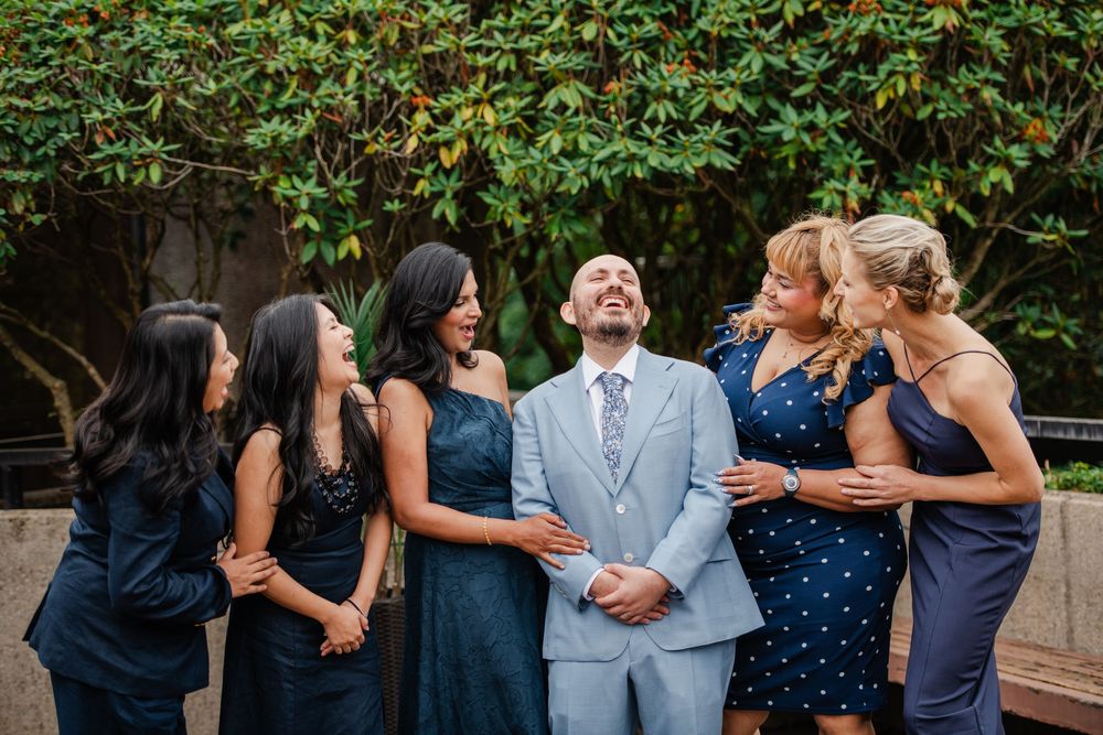 Groom in light blue suit laughing with five bridesmaids in navy dresses outdoors at a wedding celebration.