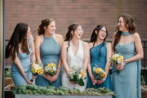 Bride in white gown laughs with four bridesmaids in blue dresses holding sunflower bouquets outdoors.