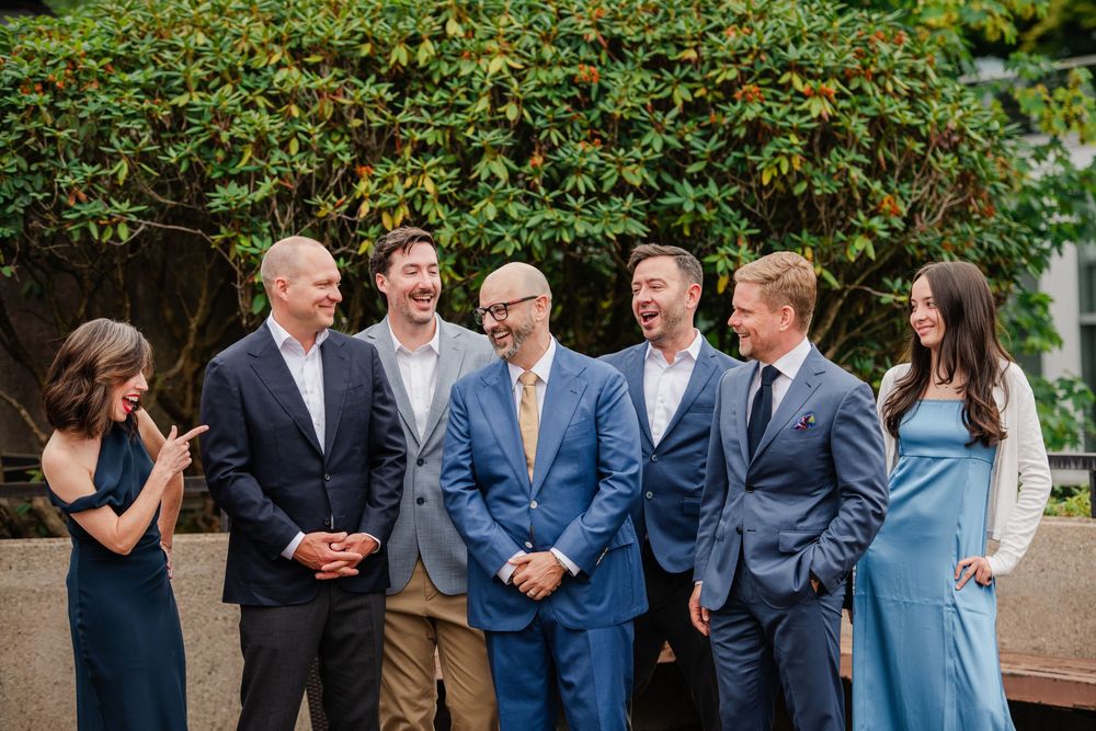 Group of elegantly dressed wedding party members laughing together outdoors near lush green foliage.