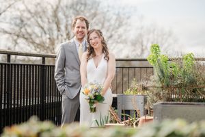 A smiling bride and groom pose together on an outdoor terrace surrounded by greenery on their wedding day.