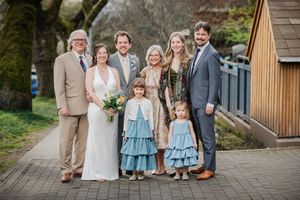 Family wedding portrait with bride, groom, flower girls in blue dresses, and relatives posing outdoors on a paved path.
