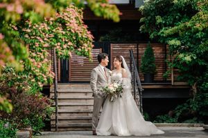 A couple in wedding attire stand together on wooden steps surrounded by lush greenery and blooming flowers.