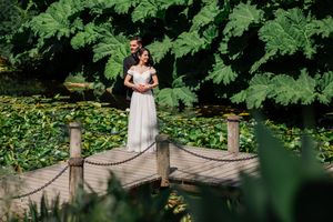 A flowing white dress stands elegantly on a wooden dock surrounded by lush greenery and a serene pond.