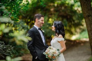 A well-dressed bride and groom share a happy moment together in a lush garden setting while holding a white floral bouquet.