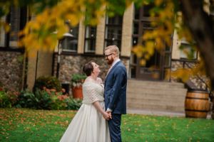 A couple embraces on a lawn with autumn leaves and rustic barrels in the background.